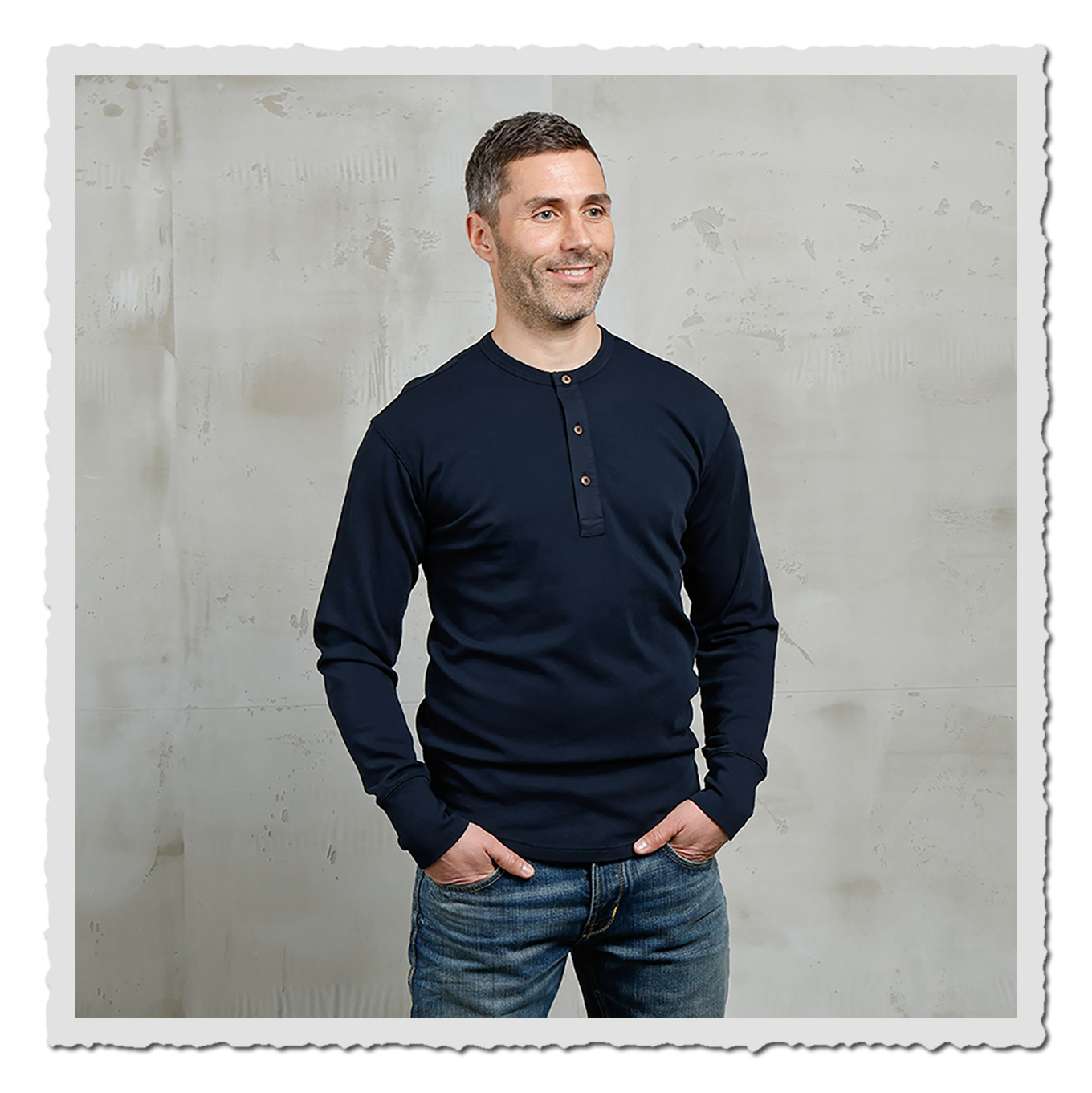 Frontansicht eines lächelnden Models im dunkelblauen Langarmshirt vor heller, strukturierter Wand.